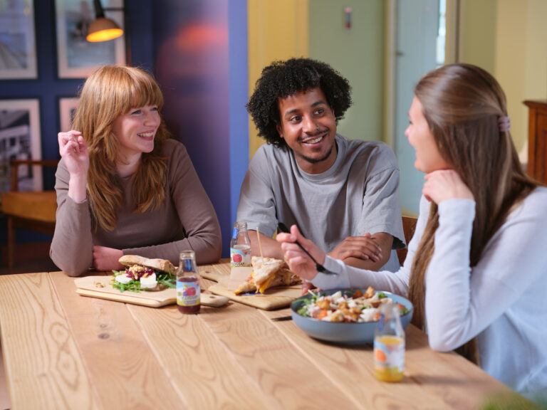 Drie mensen lunchen samen aan een houten tafel bij Jitha’s Deli met broodjes, salade en flevosap, terwijl ze lachen en praten