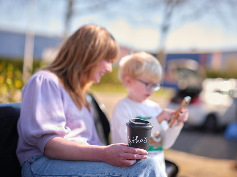 Vrouw met een Jitha’s koffiebeker in de hand zit buiten met een kind dat op een telefoon kijkt.