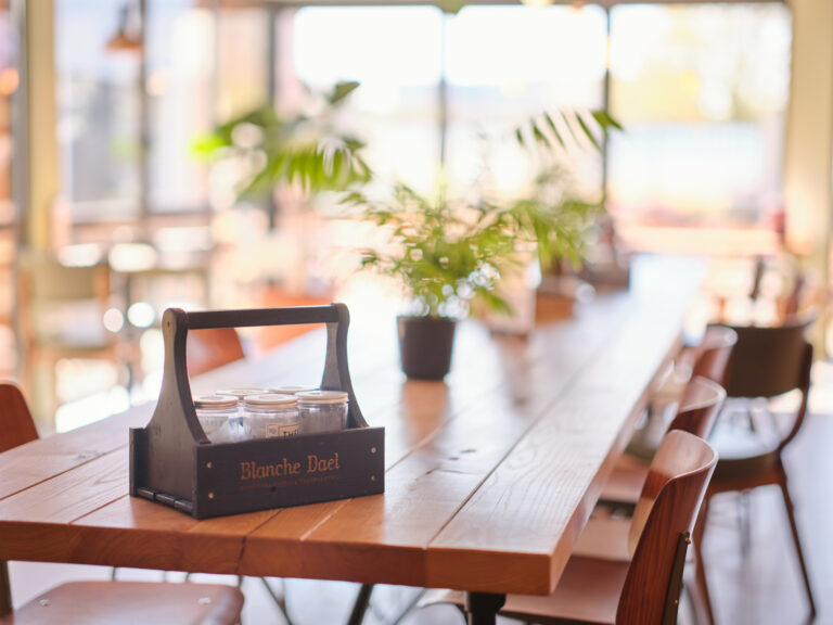 Houten stamtafel met stoelen, planten en een Blanche Dael thee in een zonnig interieur bij Jitha’s Deli