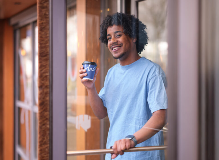 Man in blauw T-shirt glimlacht terwijl hij een beker koffie to go vasthoudt bij de ingang van Jitha’s Deli.