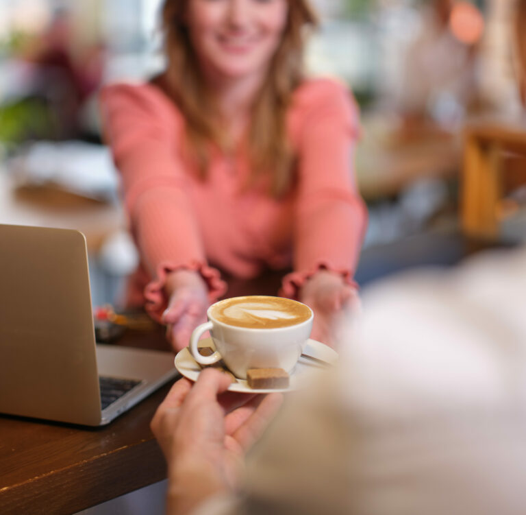 Vrouw ontvangt een vers gezette cappuccino met latte art aan tafel in een gezellige koffiezaak, met laptop op tafel bij Jitha’s Deli