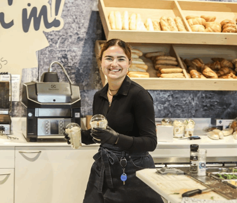 Medewerker van Smeer’m in Slagharen die glimlachend twee bakjes met vers belegde broodjes vasthoudt, met brood en koffiemachines op de achtergrond.